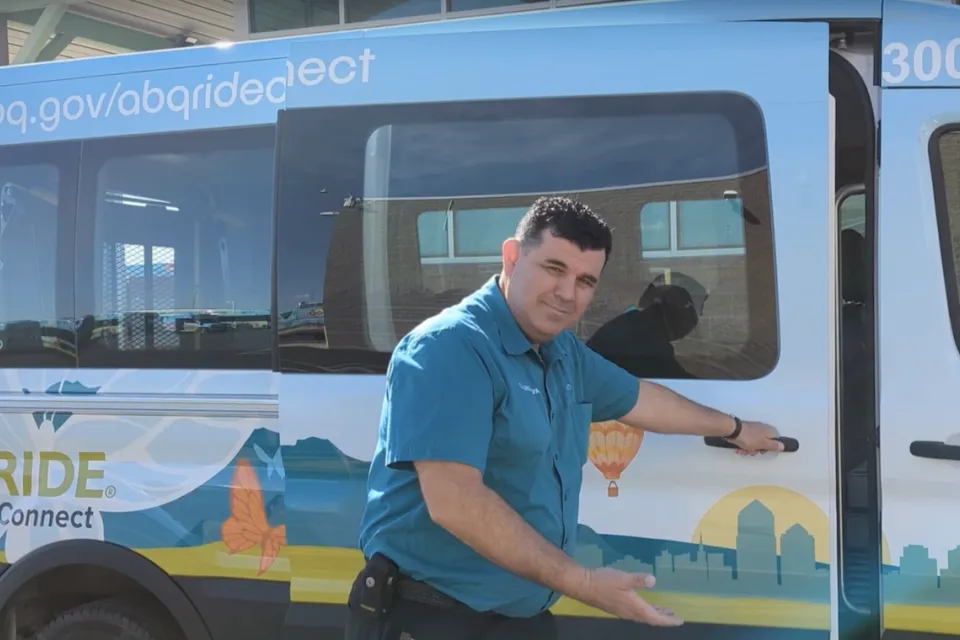 Man in teal shirt opening door to white microtransit shuttle with cactus graphics and making inviting gesture toward the camera.